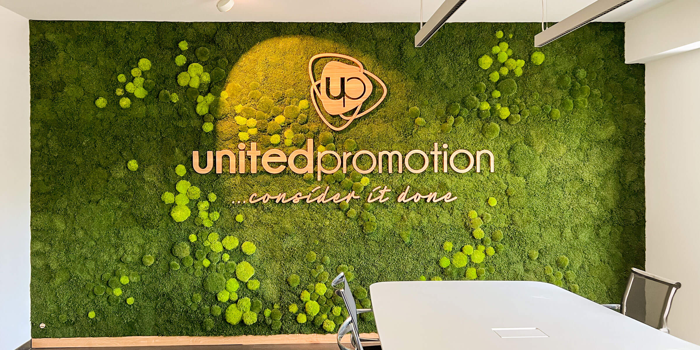 Transformation einer einfachen weißen Wand in eine künstlerische Mooswand bei United Promotion, die Nachhaltigkeit, Markenpräsentation und Natur vereint, mit einem integrierten Firmenlogo aus massiver Eiche.